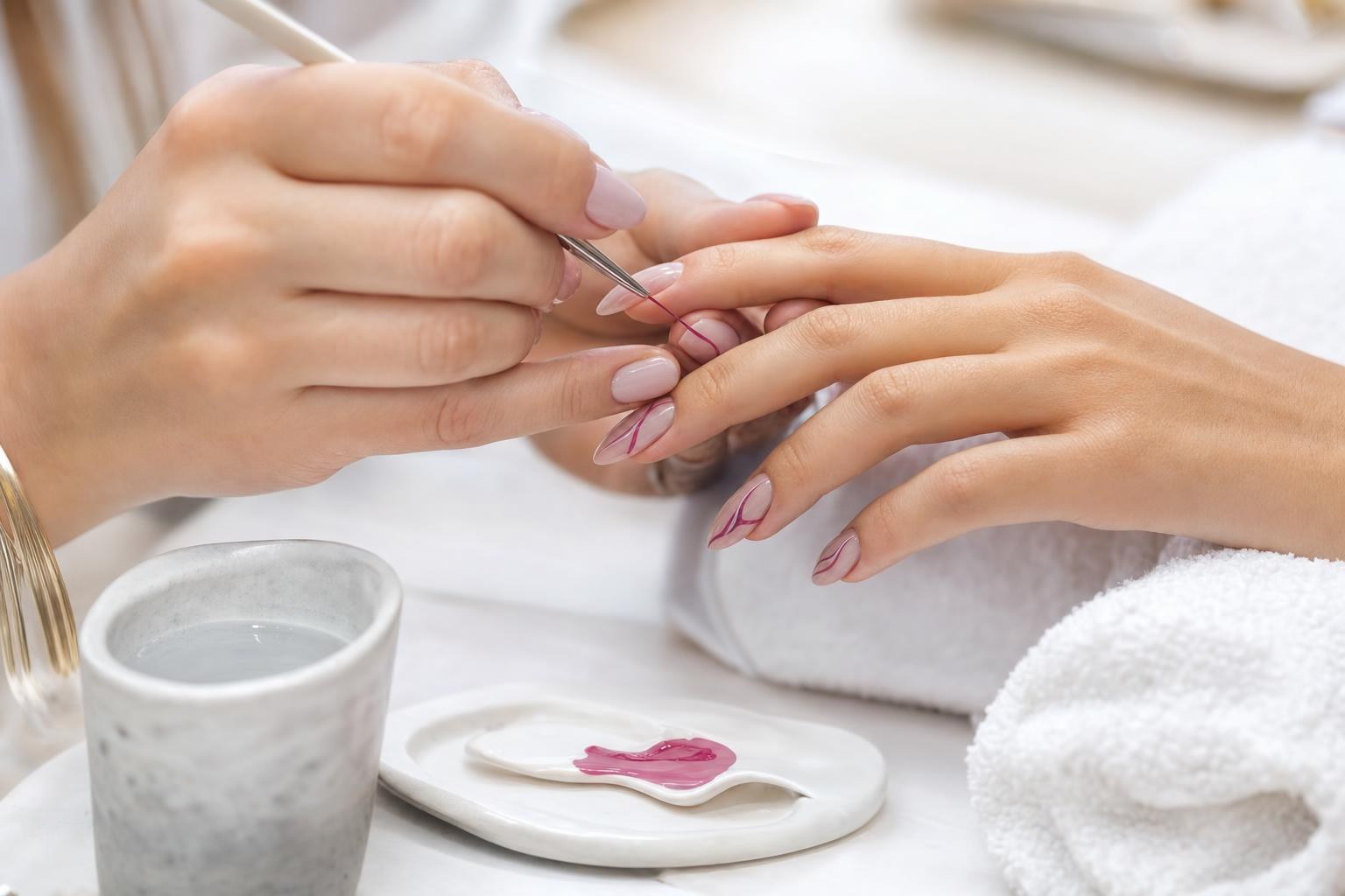 A fine brush laying a hand-painted line across an almond nail at a bright, daylit work table